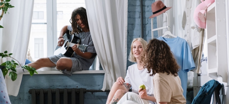 two girls and one man sitting in a dorm room with clothes hanging from the walls to depict one of the dorm storage ideas students love