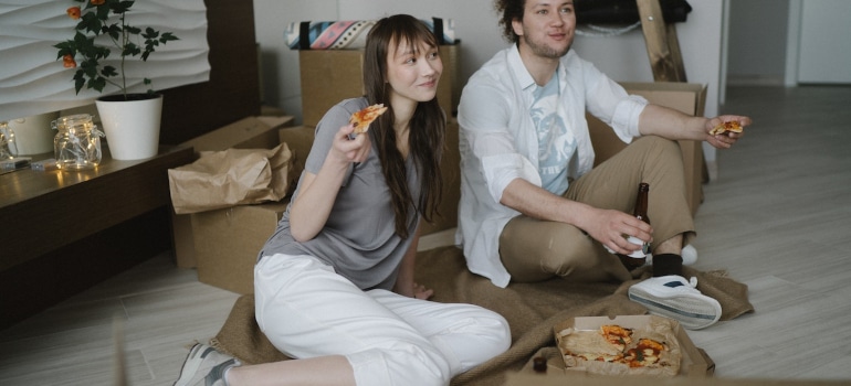 A woman and a man are sitting on the floor eating pizza and thinking about Tampa neighborhoods and home styles